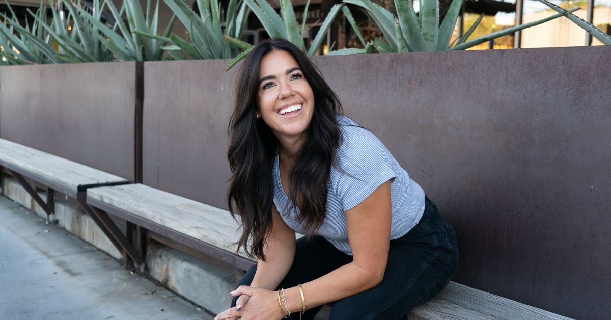 Image of a dark haired woman sitting on a bench in front of a concrete wall and smiling
